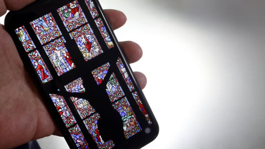 Ein Smartphone mit einem bunten Kirchenglasfenster, vor dem der Schatten einer Statue von Jesus am Kreuz abgezeichnet ist