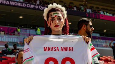 Ein Frau, die sich blutuge Tränen geschminkt hat, zeigt in einem Stadion ein Trikot mit der Aufschrift "Mahsa Amini"
