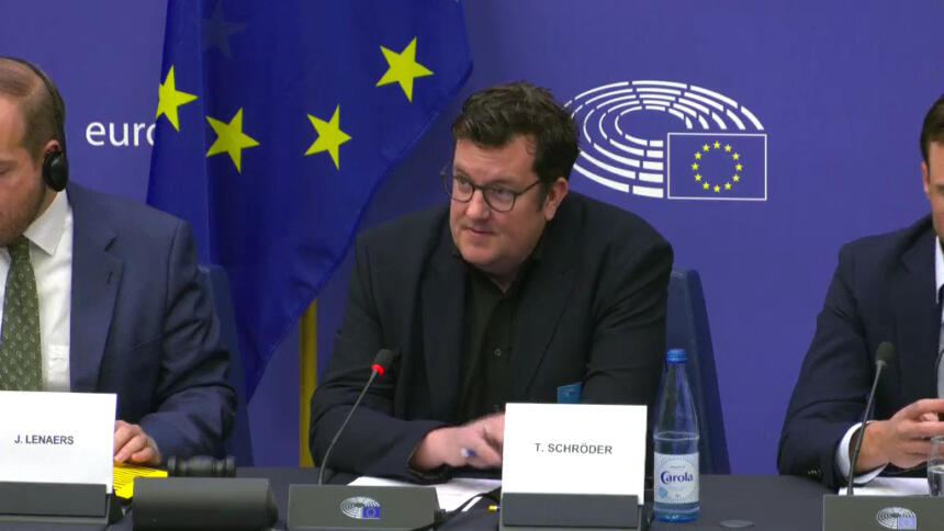 Thorsten Schröder sitzt auf dem Podium und spricht zu den Parlamentarier:innen. Im Hintergrund eine Flagge der EU, sowie das Emblem des Europäischen Parlaments.