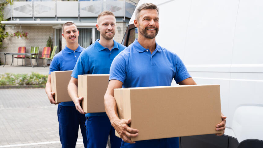 Drei wie Klone aussehende Männer in blauen T-Shirts tragen gleich große braune Kartons vor einem weißen Transporter