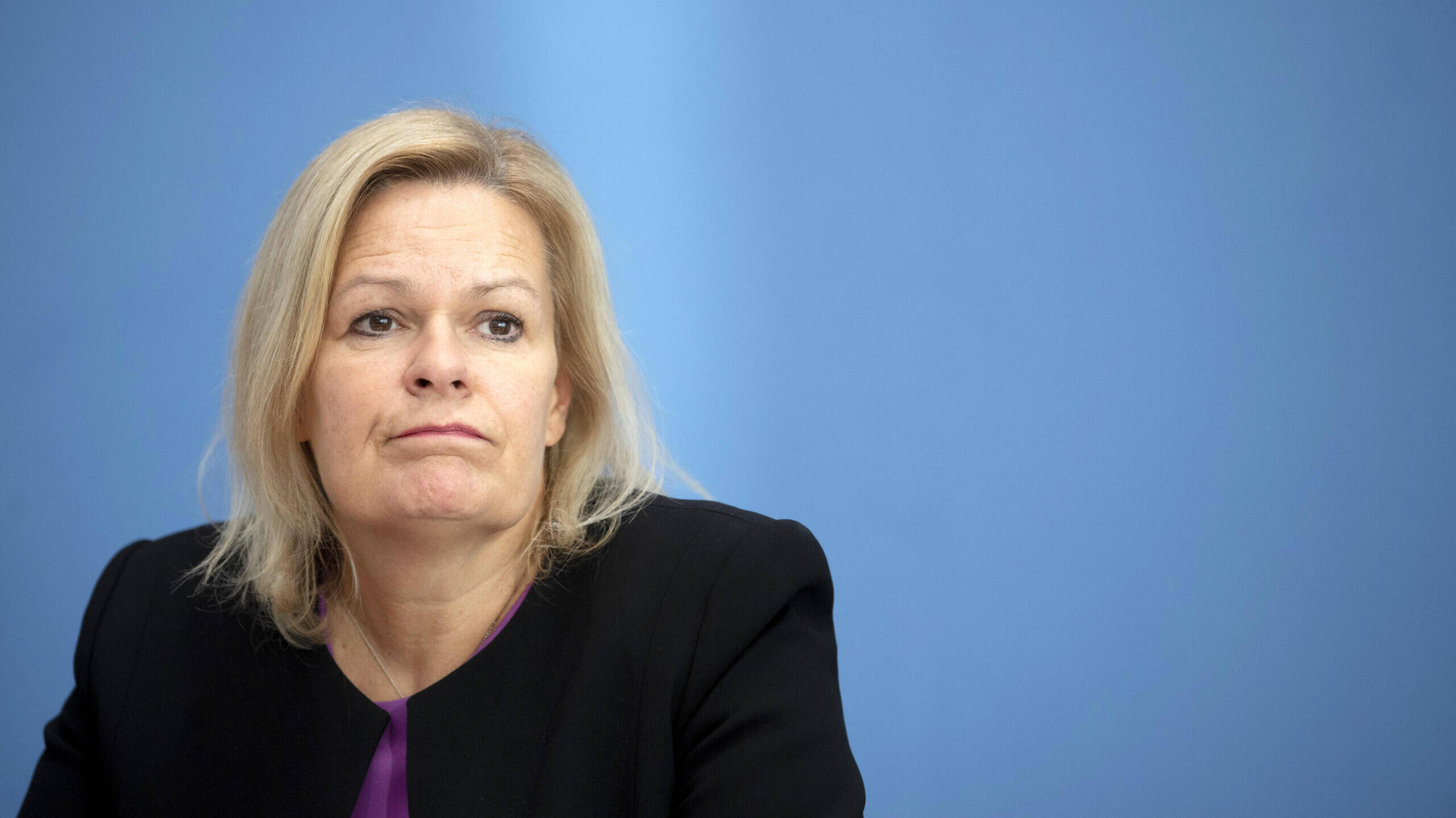 Eine blonde Frau mittleren Alters vor einer blauen Wand, ihr Gesichtsausdruck wirkt unzufrieden