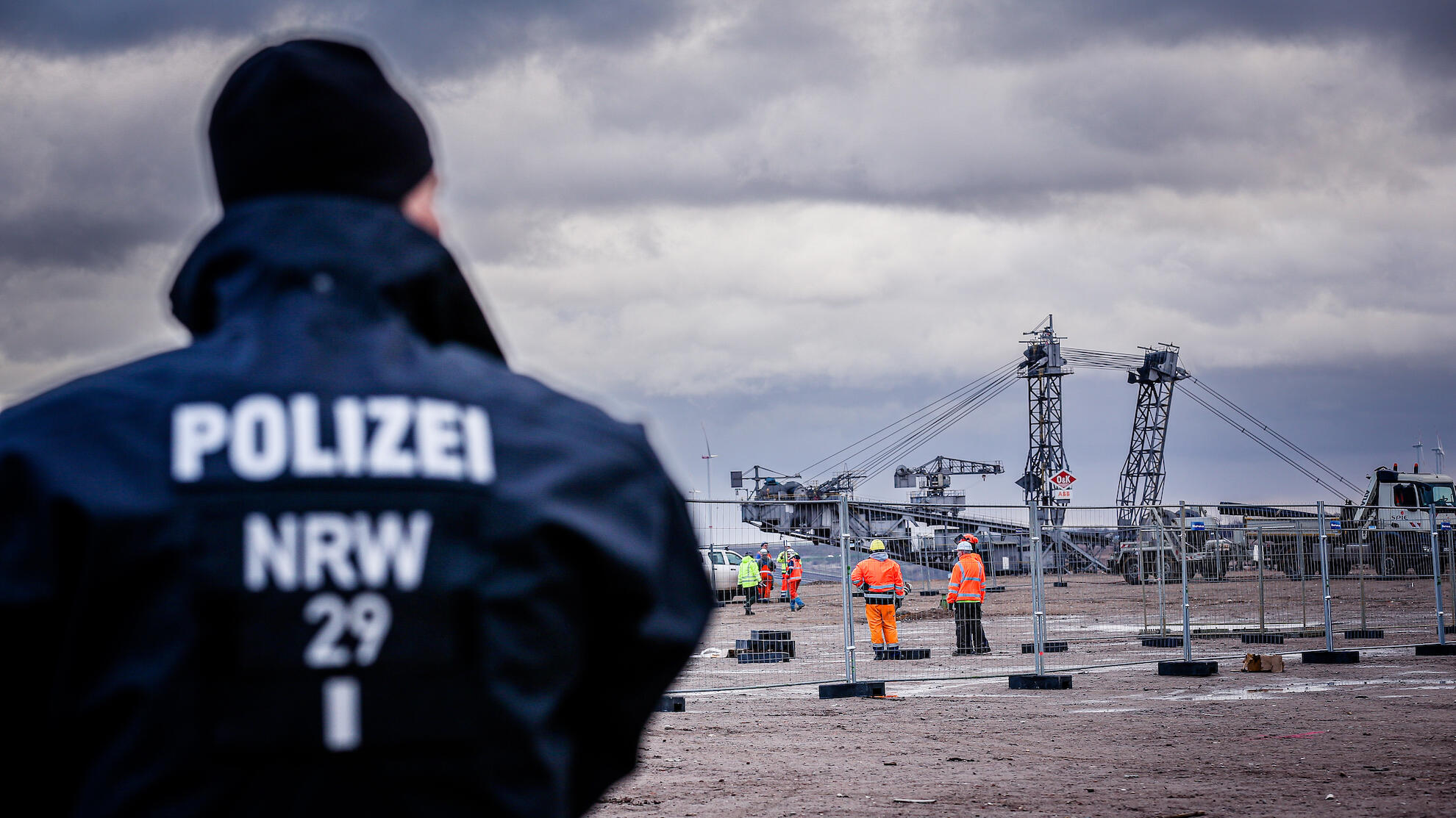 Polizist steht vor einem Bauzaun, im Hintergrund Mitarbeiter von RWE in orangenen Westen und eine Schaufelradbagger