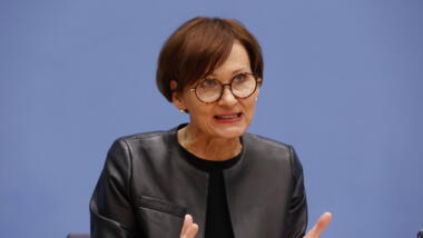 Bundesministerin für Bildung Forschung Bettina Stark-Watzinger mit schwarzer Jacke, kurzen roten Haaren und Briller vor blauem Hintergrund