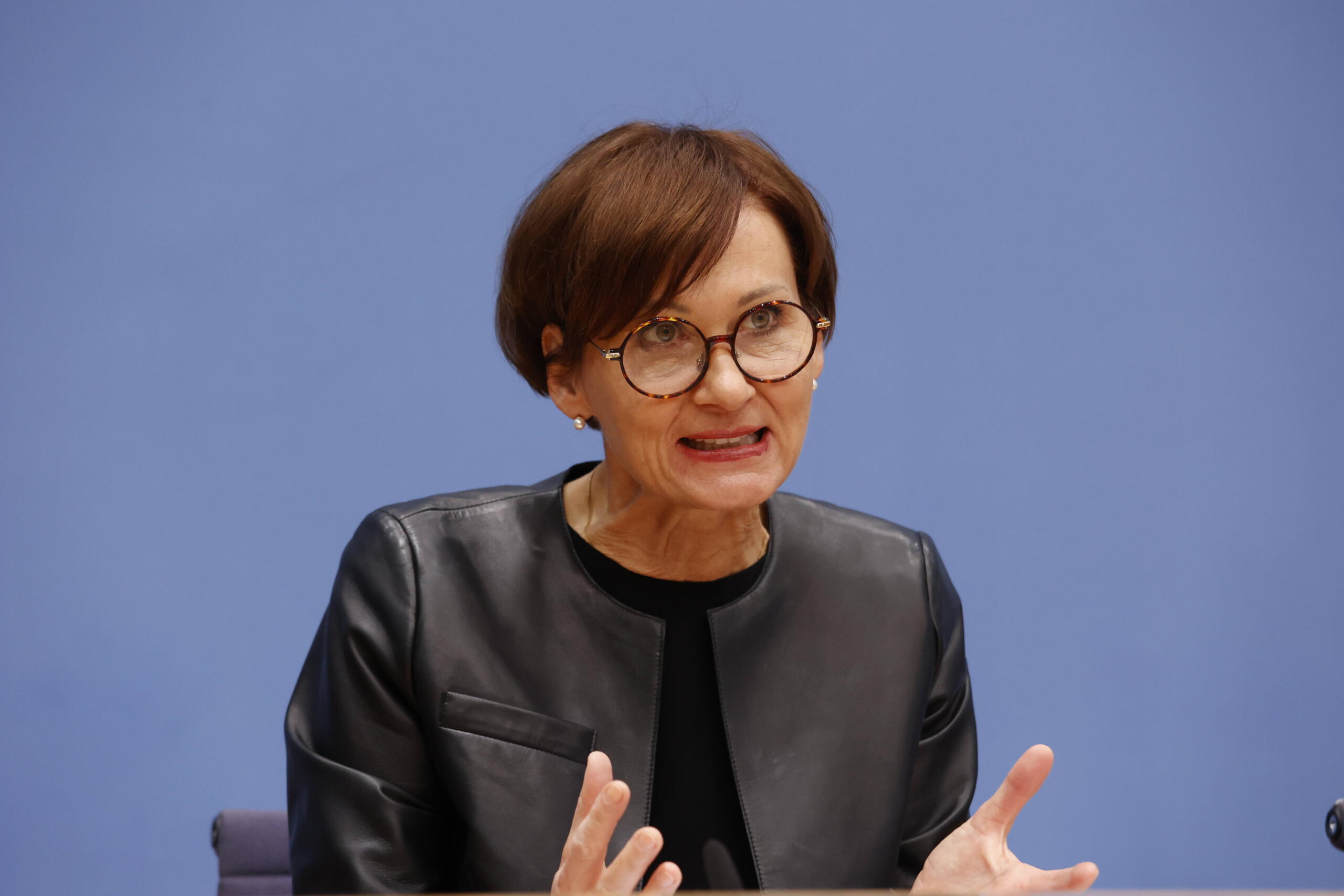 Bundesministerin für Bildung Forschung Bettina Stark-Watzinger mit schwarzer Jacke, kurzen roten Haaren und Briller vor blauem Hintergrund