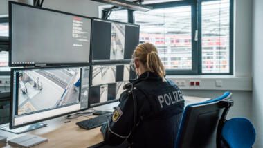 Eine Polizistin sitzt vor mehreren Bildschirmen, die Szenen am Bahnhof aufnehmen.