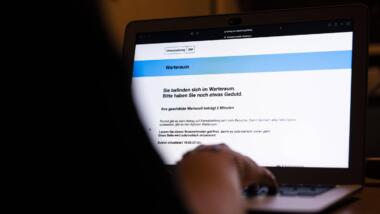 Mensch vor einem Laptop, auf dem Bildschirm ist die Webseite einmalzahlung200.de zu sehen; dem Menschen wird angezeigt, er befinde sich im digitalen Warteraum