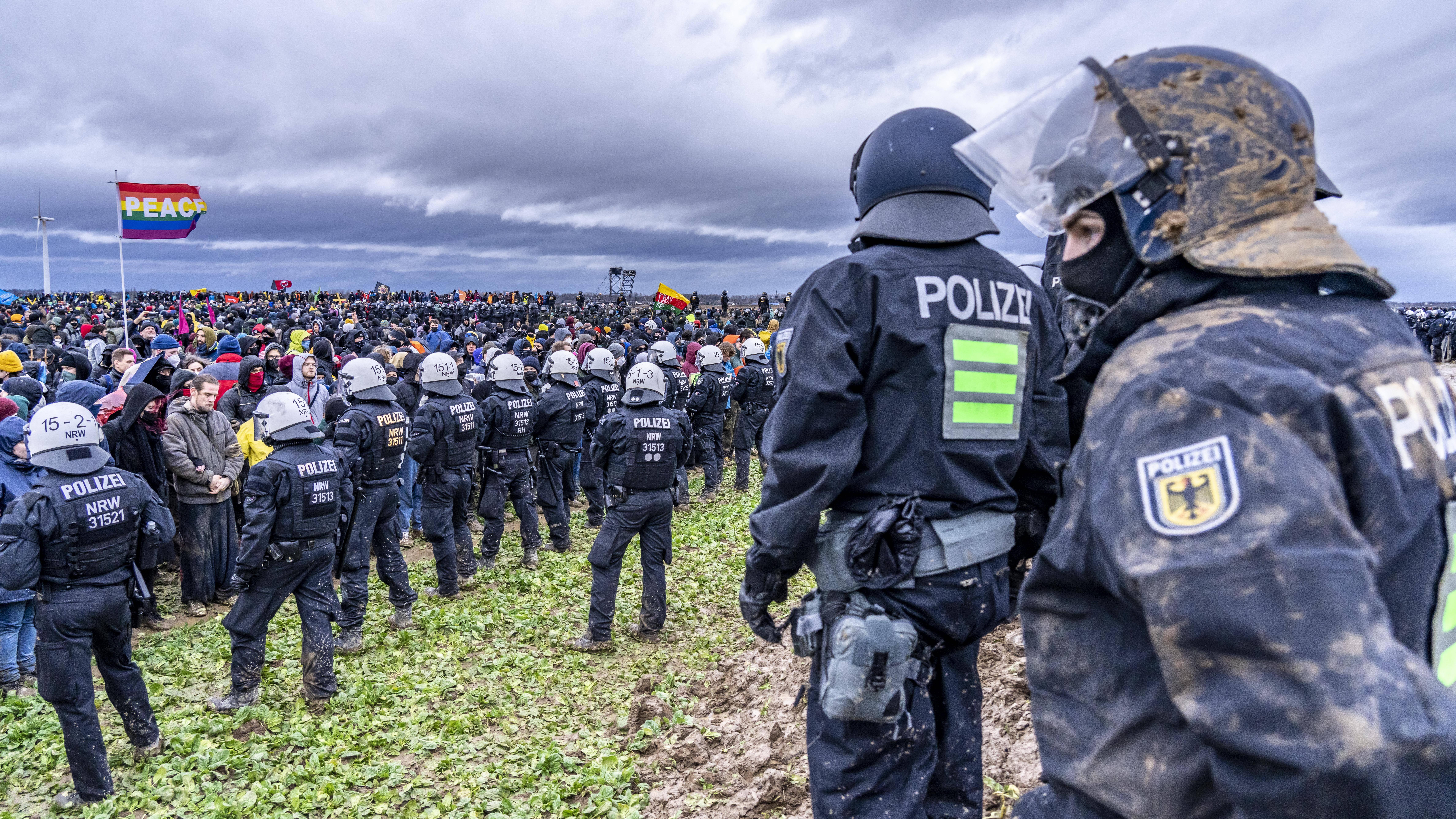 Eine Polizeikette steht Demonstrierenden entgegen. Es weht eine Pace-Fahne.