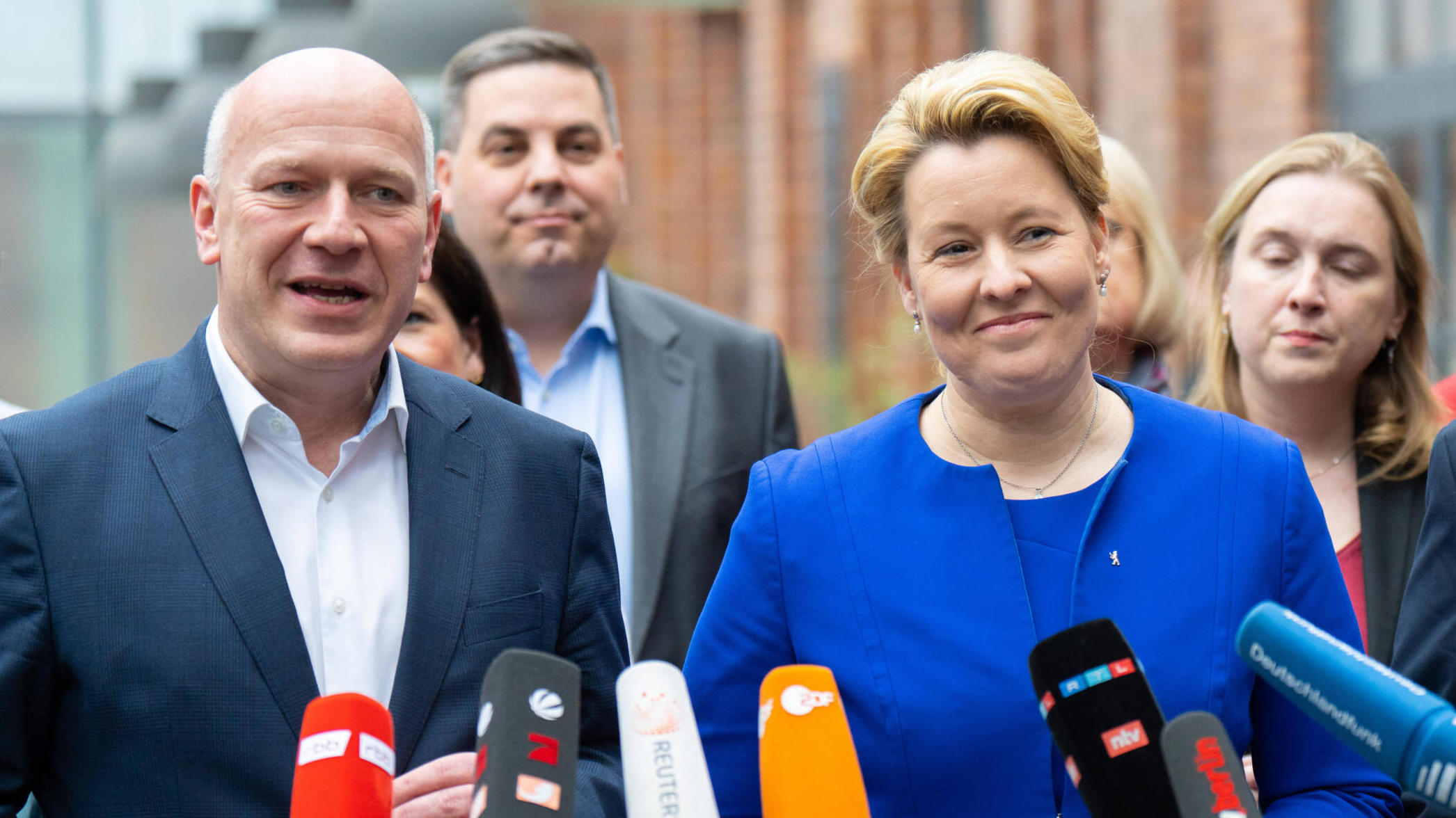 Ganz im Vordergrund Mikrofone, dahinter zwei Menschen in blauen Anzügen, dahinter weitere Menschen