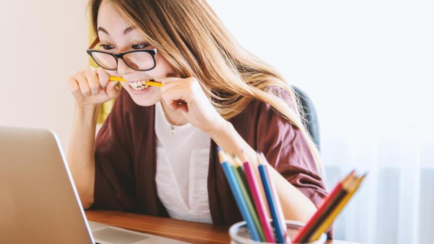 Eine junge Person mit langen Haaren und Brille sitzt vor dem Laptop und kaut mit verzwiefeltem Gesichtsausdruck auf einem Bleistift