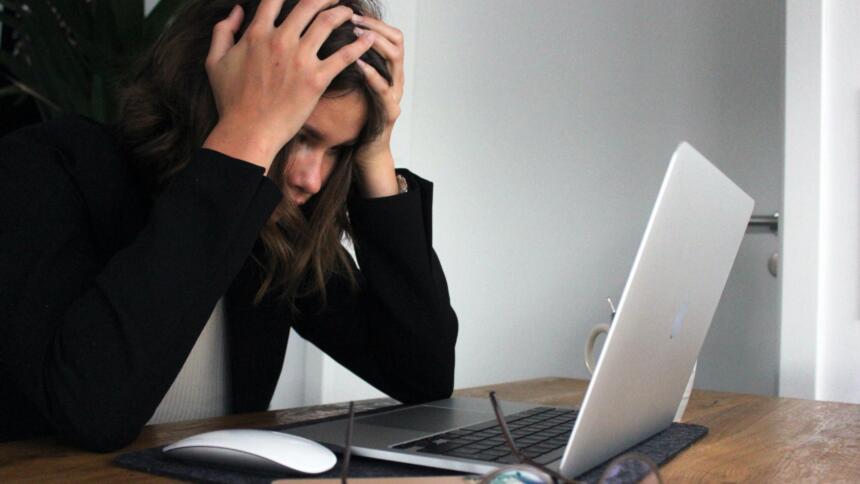 Eine Person lin kangen, dunklen Haaren sitzt vor dem Laptop und fasst sich mit beiden Händen an den Kopf