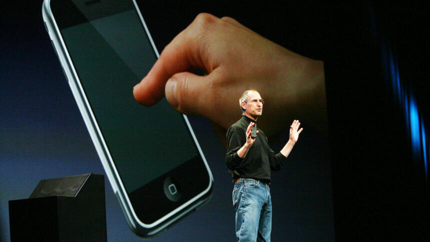 Steve Jobs im Jahr 2007 vor einer großen Leinwand, auf der das erste iPhone zu sehen ist