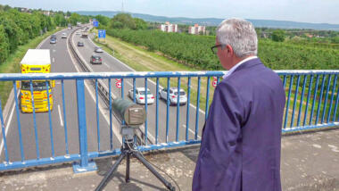 Innenminister schaut von Brücke auf Autobahn, vor ihm eine Kamera.