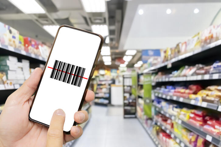 Im Vordergrund eine Hand, die ein Smartphone hält auf dem ein Barcode angezeigt wird, im Hintergrund ein Supermaktregal