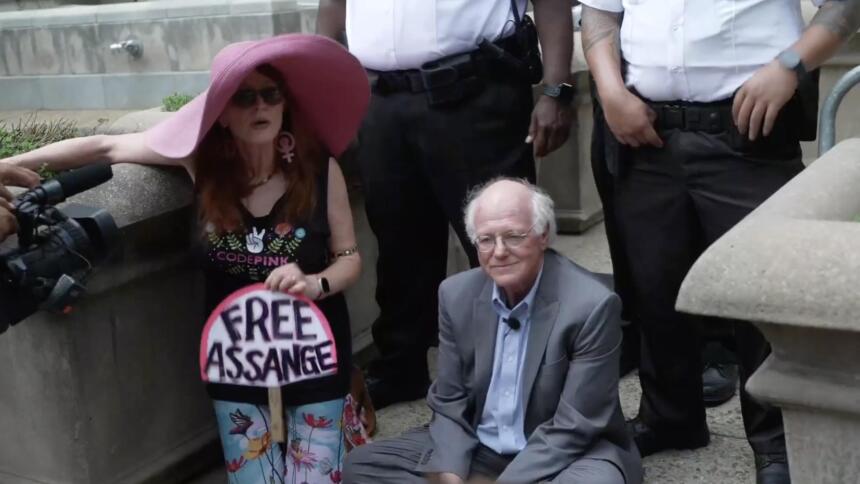 Jodie Evans mit einem Schild "Free Assange" neben dem sitzenden Ben Cohen.