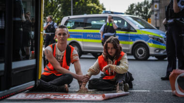 Zwei Aktivist:innen der Letzten Generation sitzen auf der Straße, dahinter ein Streifenwagen