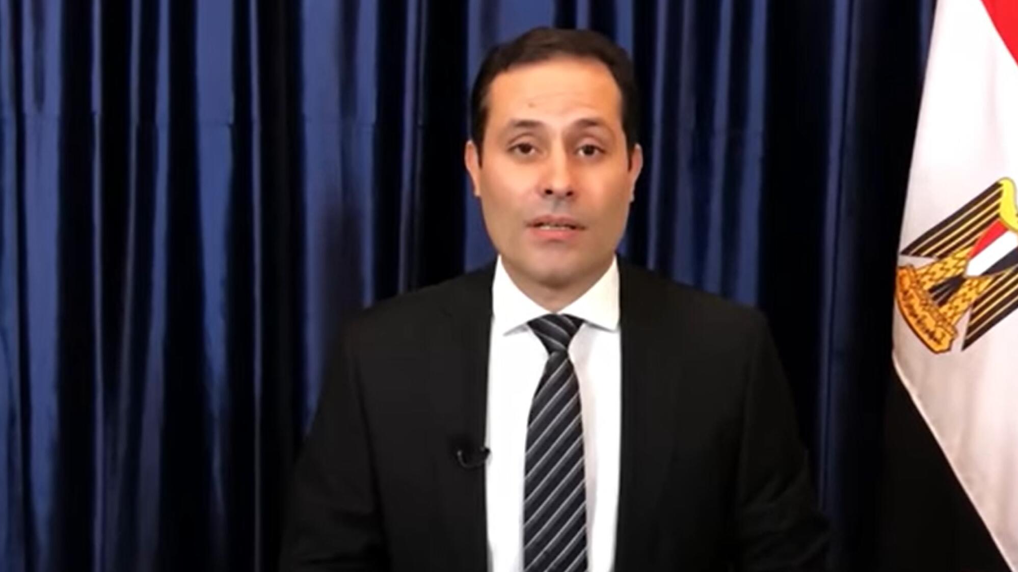 Ein Mann im Anzug vor einem blauen Vorhang und einer Flagge