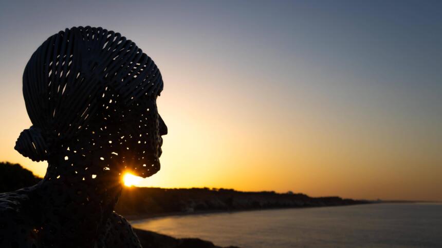 Eine Metall-Skulptur des Künstlers Carlos de Oliveira Correia blickt auf der Aussichtsplattform am Strand von Olhos des Augua bei Sonnenaufgang auf den Atlantik hinaus.