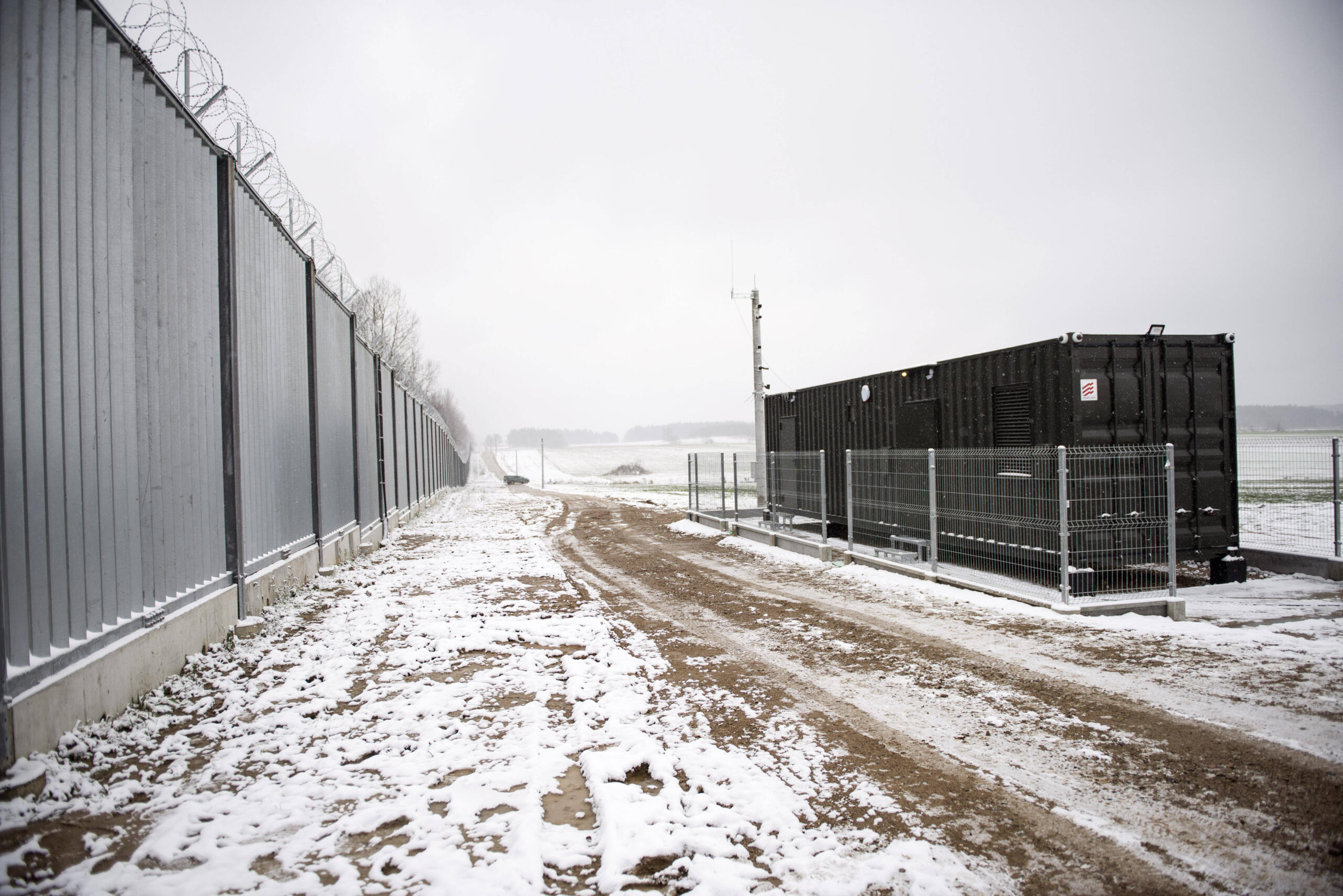 Links hoher Metallzaun, davor verschneiter Boden, rechts ein schwarzer Metallcontainer