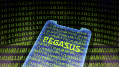 Ein Smartphone, auf dessen Bildschirm in neongrünen Buchstaben "Pegasus" steht. Im Hintergrund ist Binärcode zu sehen.