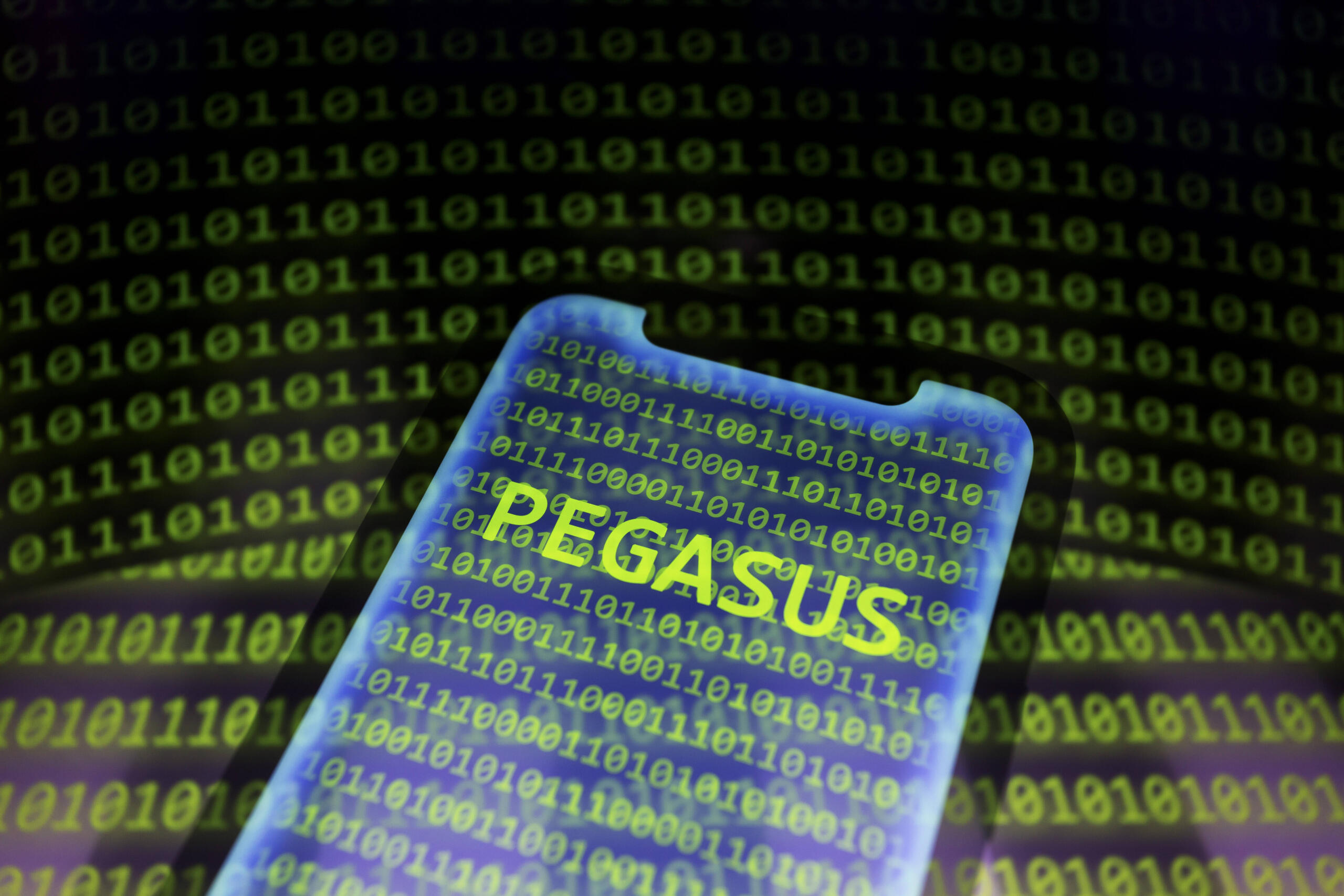 Ein Smartphone, auf dessen Bildschirm in neongrünen Buchstaben "Pegasus" steht. Im Hintergrund ist Binärcode zu sehen.