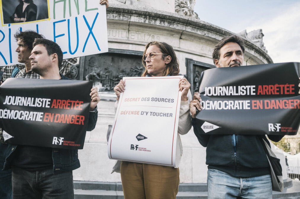 Drei Menschen, die bei einer Demonstration vor einem Brunnen stehe und Schilder hochhalten. Auf einem steht auf französisch "Journalisten verhaftet, Demokratie in Gefahr"