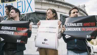 Drei Menschen, die bei einer Demonstration vor einem Brunnen stehe und Schilder hochhalten. Auf einem steht auf französisch "Journalisten verhaftet, Demokratie in Gefahr"
