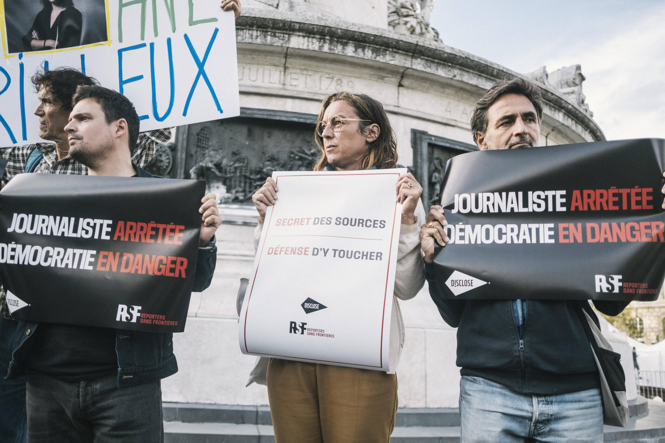 Drei Menschen, die bei einer Demonstration vor einem Brunnen stehe und Schilder hochhalten. Auf einem steht auf französisch "Journalisten verhaftet, Demokratie in Gefahr"