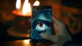 Eine Person hält ein Smartphone, aud dem ein spionierendes, rotes Auge abgebildet ist