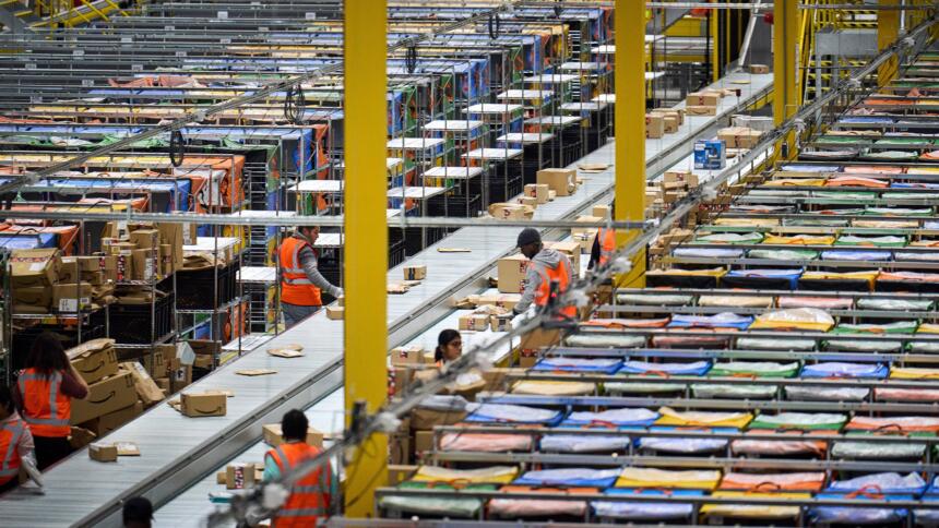 Das Bild zeigt das innere eines Amazon-Lagerhauses. Arbeiter in orangen Westen legen Pakete auf Fließbänder.