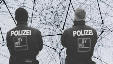 Zwei Polizisten, mit dem Rücken zur Kamera, die auf eine komplexe Linienstruktur schauen. Grautöne.