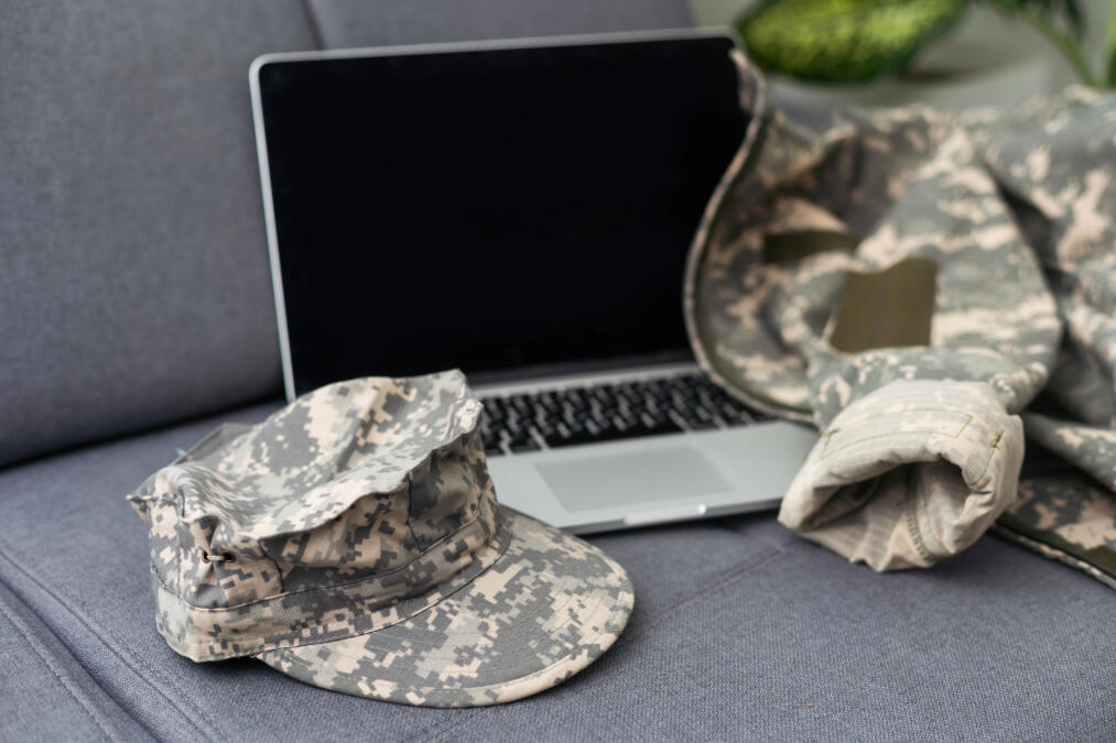 Ein Laptopn, davor eine Militärmütze in Camouflage und dahinter eine Jacke in Camouflage