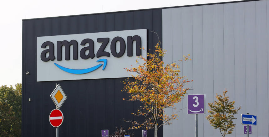 Eine Lagerhalle mit einem riesigen Amazon-Logo, davor Verkehrsschilder und Bäume.