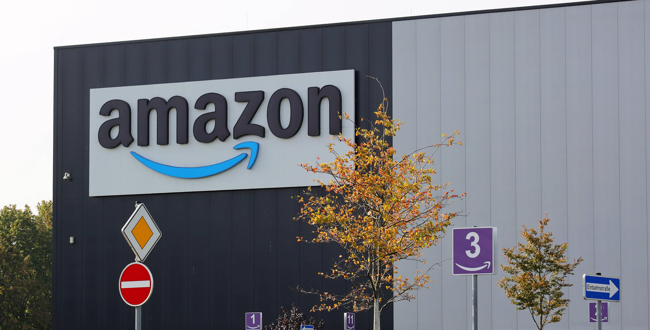 Eine Lagerhalle mit einem riesigen Amazon-Logo, davor Verkehrsschilder und Bäume.