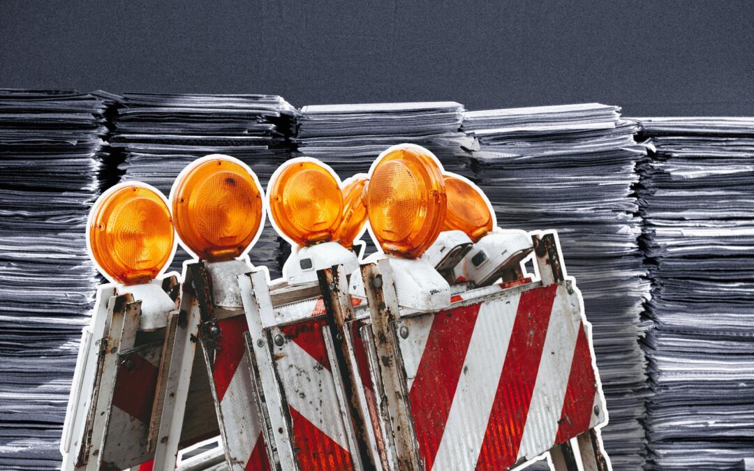 Baustellenabsperrung mit Warnleuchten, im Hintergrund Aktenstapel dicht an dicht