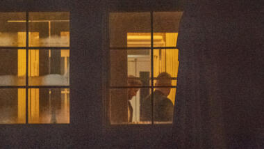 Blick von außen auf ein erleuchtetes Fenster, innen sind zwei Personen zu sehen.
