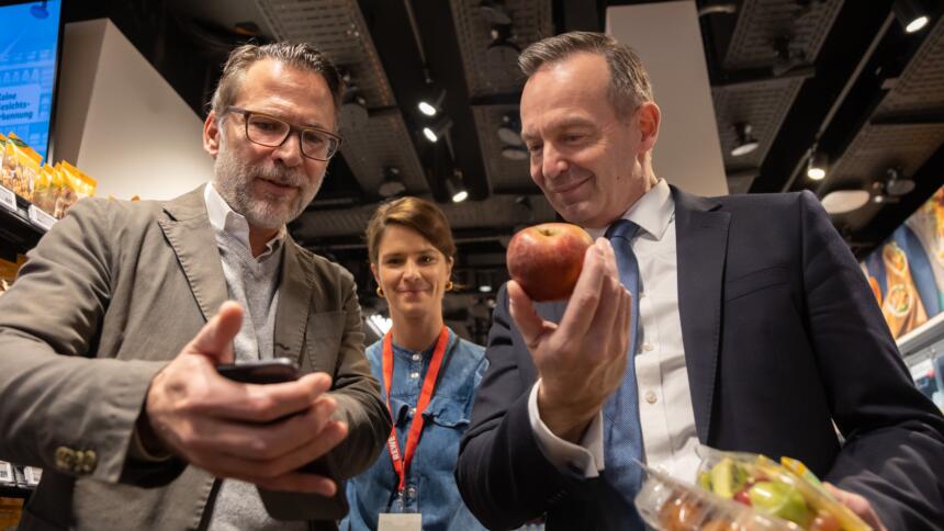 Zwei Männer mit Anzügen, im Hintergrund eine Frau. Der Mann links schaut auf ein Smartphone, der Mann rechts (Wissing) hält lachend einen Apfel hoch.