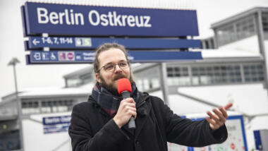 Mann mit langen Haaren und Brille steht vor einem Schild mit der Aufschrift "Berlin Ostkreuz" und hält ein rotes Mikrofon in der Hand, in das er spricht, währen er mit der anderen Hand eine ausladende Geste macht.