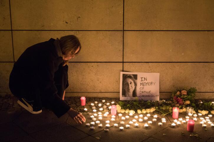 Eine Frau kniet vor einer Mauer und zündet eine Kerze an. Umgeben von Kerzen ist ein Bild von Daphen Caruana Galizia zu sehen.