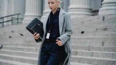 Eine junge Frau im Mantel mit Laptop unter dem Arm geht mit unglücklichem Gesicht eine steinerne Treppe hinunter, die zu einem offiziellen Gebäude zu gehören scheint.