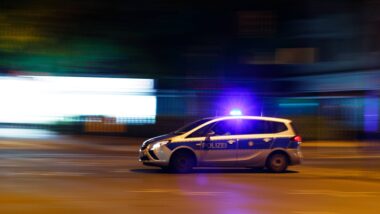 Abgebildet ist Polizeiwagen mit angeschaltetem Blaulicht im Profil. Es ist Nacht, im Hintergrund sind Leuchtreklamen zu sehen. Die Bewegungsunschärfe erweckt den Eindruck, der Wagen fahre schnell.
