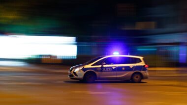 Abgebildet ist Polizeiwagen mit angeschaltetem Blaulicht im Profil. Es ist Nacht, im Hintergrund sind Leuchtreklamen zu sehen. Die Bewegungsunschärfe erweckt den Eindruck, der Wagen fahre schnell.