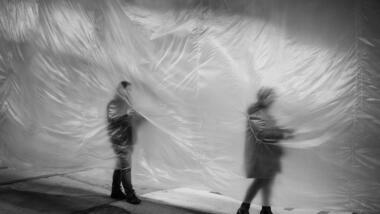 Auf dem schwarz-weißen Bild sind zwei Menschen zu sehen, die sich ihren Weg entlang einer halbdurchsichtigen Folie bahnen.