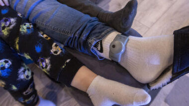 Abgebildet ist das Bein einer Person, die einen weißen Socken und eine elektronische Fußfessel trägt. Links und rechts davon ist je ein Bein und Fuß eines Kindes zu sehen.