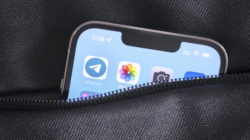 Ein Smartphone steckt in einer Tasche mit Reißverschluss. Lediglich die obere Ecke schaut raus. Auf dem Display ist das Logo der App "Telegram" zu sehen, daneben die Logos für die Apps "Photos" und "Camera".