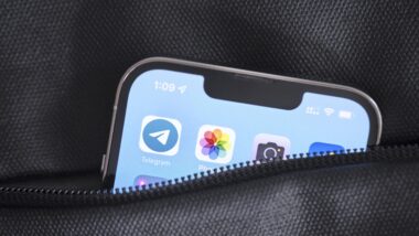 Ein Smartphone steckt in einer Tasche mit Reißverschluss. Lediglich die obere Ecke schaut raus. Auf dem Display ist das Logo der App "Telegram" zu sehen, daneben die Logos für die Apps "Photos" und "Camera".
