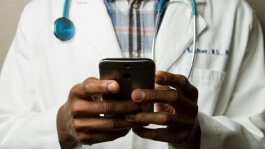 Eine Person, deren Kopf nicht zu sehen ist, hält ein Smartphone in beiden Händen. Sie trägt einen Arztkittel und ein Stethoskop.