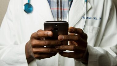 Eine Person, deren Kopf nicht zu sehen ist, hält ein Smartphone in beiden Händen. Sie trägt einen Arztkittel und ein Stethoskop.