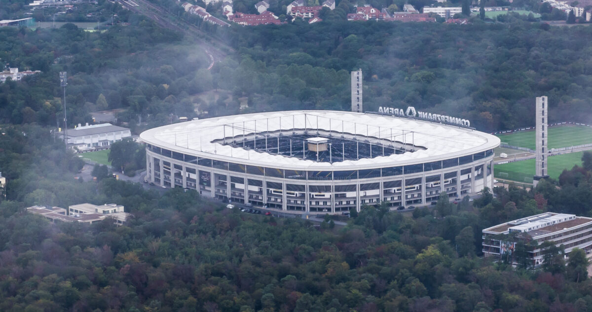Zu sehen ist die Frankfurter Arena. Das Stadion ist umgeben von vielen Bäumen und einigen kleineren Gebäuden. Es ist etwas neblig.