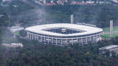 Zu sehen ist die Frankfurter Arena. Das Stadion ist umgeben von vielen Bäumen und einigen kleineren Gebäuden. Es ist etwas neblig.