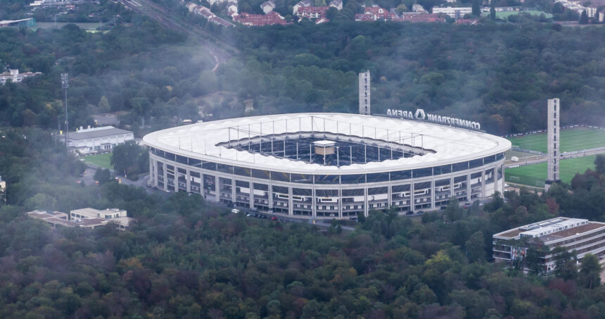 Zu sehen ist die Frankfurter Arena. Das Stadion ist umgeben von vielen Bäumen und einigen kleineren Gebäuden. Es ist etwas neblig.
